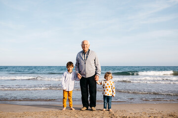 Portrait of happy grandfather standing hand in hand on the beach with his grandchildren