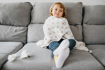 Portrait of sick little girl sitting on the couch at home wrapped in blanket
