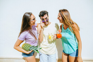 Happy friends holding refreshing drinks and pineapple in front of white wall