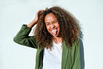 Playful woman sticking out tongue against white wall during sunny day