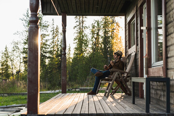 Young man sitting on veranda of a wood house, playing the ukulele