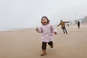 Happy little girl with lolly running on the beach in winter while her brother playing in the...