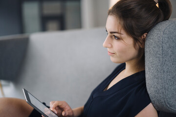 Young businesswoman sitting on couch with digital tablet looking at distance