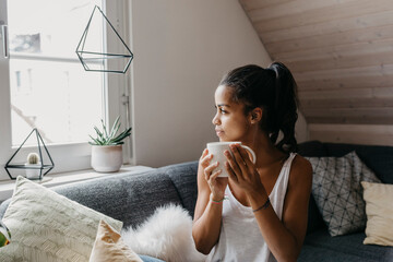 Young woman with cup of coffee sitting on the couch at home looking out of window - obrazy, fototapety, plakaty