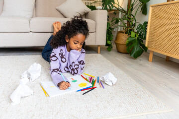 POC girl lying on carpet infront of sofa coloring with crayons