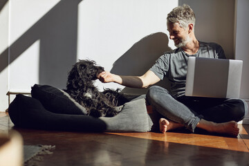 Mature man sitting on the floor at home with laptop and dog