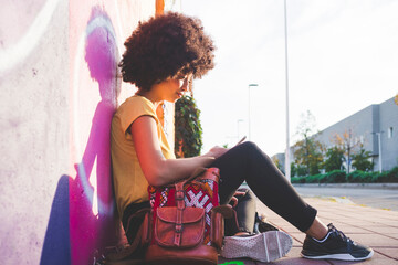 Young woman with afro hairdo sitting at graffiti wall and using smartphone