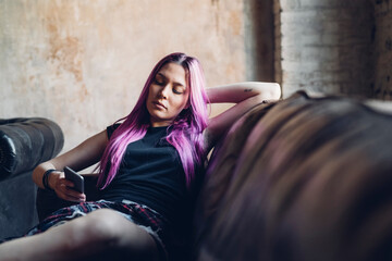 Young woman with pink hair sitting on sofa in a loft using smartphone