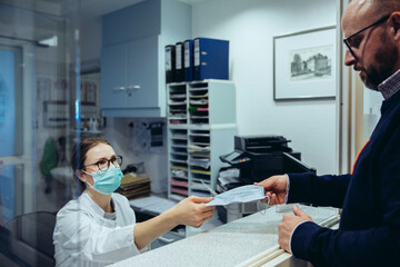 Employee at reception desk of hospital ward handing over mask to visitor - obrazy, fototapety, plakaty