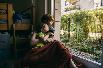 Mother and little son sitting on the floor at home looking out of window
