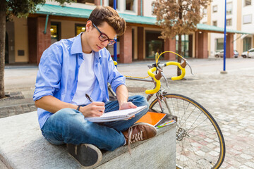 Young man with racing cycle sitting on bench writing on notepad