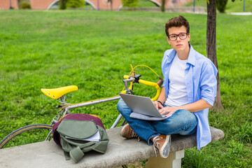 Pensive young man with racing cycle sitting on a bench using laptop