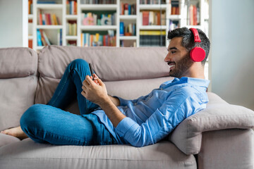 Smiling young man lying on the couch at home using smartphone and wireless headphones - Powered by Adobe
