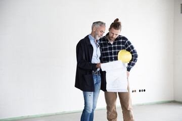 Architect and construction worker discussing blueprint while standing in empty house