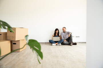 Man sitting with wife using laptop on hardwood floor against wall in new unfurnished home
