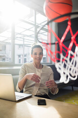 Portrait of smiling freelancer sitting at desk in a loft throwing basketball into hoop