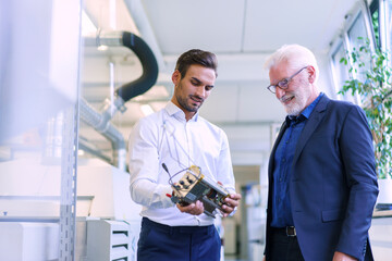 Young male engineer holding machine part while discussing with smiling senior manager at factory
