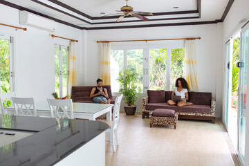 Thailand, couple sitting in spacious living room at holiday resort using electronic devices - obrazy, fototapety, plakaty