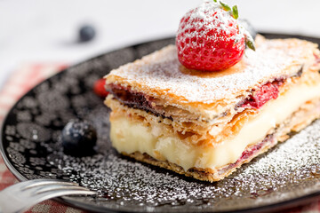 A piece of Napoleon cake with poppy seeds and cherries, decorated with strawberries, blueberries on a light background. Close up