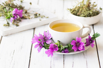 Cup of common mallow tea with fresh blooming malva sylvestris plant on white rustic table,...