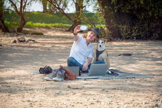Smiling woman sitting on blanket on beach with dog taking a selfie
