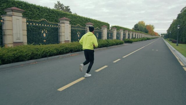 Young sportsman wearing neon green sports jacket, black trousers and bini hat running on a gloomy day passing by hedges of evergreen trees. Autumn. High quality 4k footage