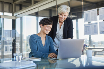 Two smiling businesswomen sharing laptop at desk in office - obrazy, fototapety, plakaty