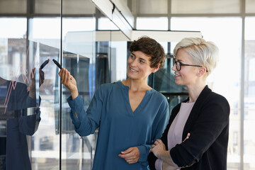 Two smiling businesswomen looking at chart on glass pane in office