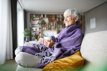 Smiling senior woman with blanket holding coffee cup while sitting on sofa at home