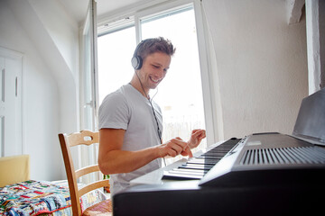 Smiling young man at home wearing headphones playing digital piano