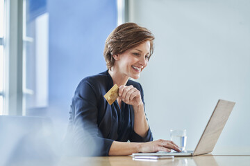 Smiling businesswoman holding credit card and using laptop at desk in office