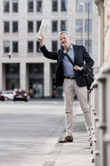 Grey-haired businessman with newspaper calling a taxi in the city - obrazy, fototapety, plakaty
