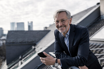 Grey-haired businessman on balcony holding smartphone smiling into camera