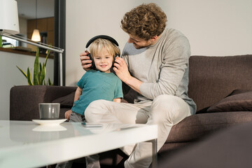 Father putting on headphones on son on couch at home