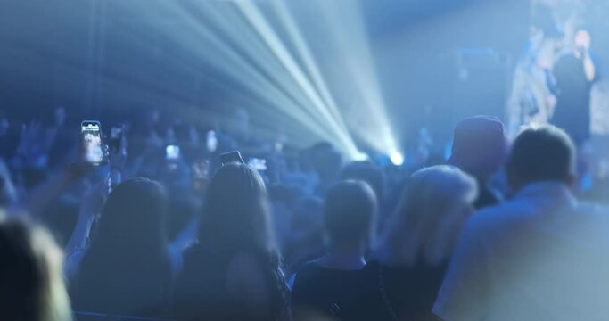 Fans crowd listen music group, watch at stage performance, back view. Happy people dance in blue bright light of professional spotlights. Audience shoot stories, video, photo on phones music concert