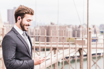 USA, New York City, smiling businessman with cell phone and earbuds on Brooklyn Bridge