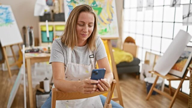 Attractive blonde artist confidently texting, smiling at her canvas in a bustling art academy studio