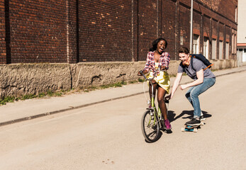 Young woman on bicycle pulling young man, standing on skateboard