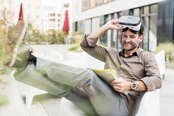 Smiling man with Virtual Reality Glasses and documents sitting on terrace