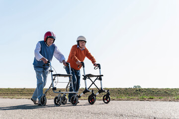 Two old friends wearing safety helmets, competing in a wheeled walker race - obrazy, fototapety, plakaty