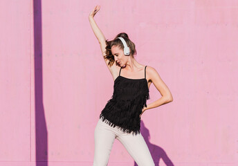 Happy young woman wearing headphones dancing in front of pink wall