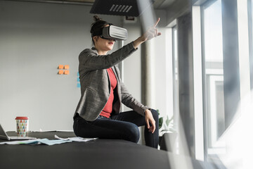 Businesswoman sitting on desk in office wearing VR glasses