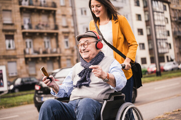 Laughing young woman pushing happy senior man with headphones and smartphone in wheelchair