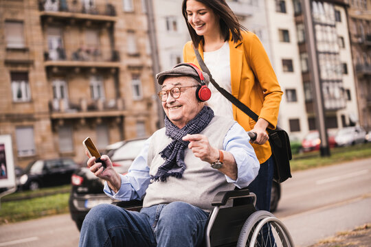 Laughing young woman pushing happy senior man with headphones and smartphone in wheelchair