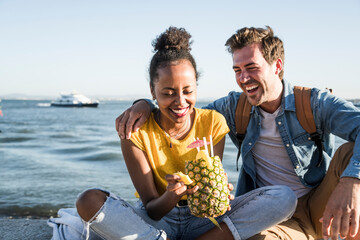 Happy young couple sitting on pier at the waterfront with a pineapple, Lisbon, Portugal - obrazy, fototapety, plakaty