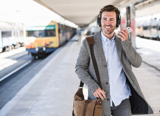 Smiling young businessman with cell phone and headphones at the train station