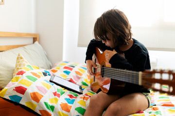 Boy sitting on bed using digital tablet for playing song on guitar