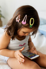 Little girl with three hair clips sitting on bed looking at smartphone