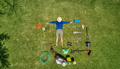 View from above of a gardener with sun hat on his face, laying on the grass with all the tools he...