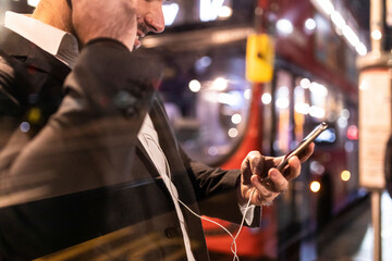UK, London, businessman with cell phone and earbuds at the bus station by night